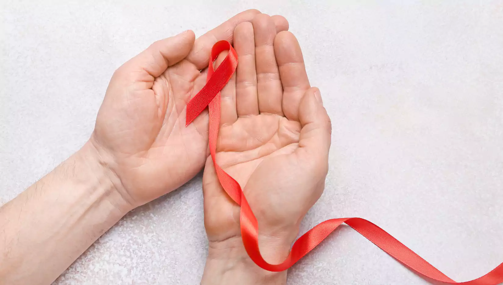 Male hands with red ribbon on light background, closeup. Cancer aids awareness concept
