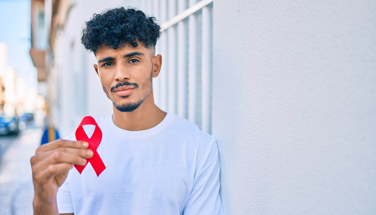 Young arab man with serious expression holding hiv awaraness red ribbon leaning on the wall.