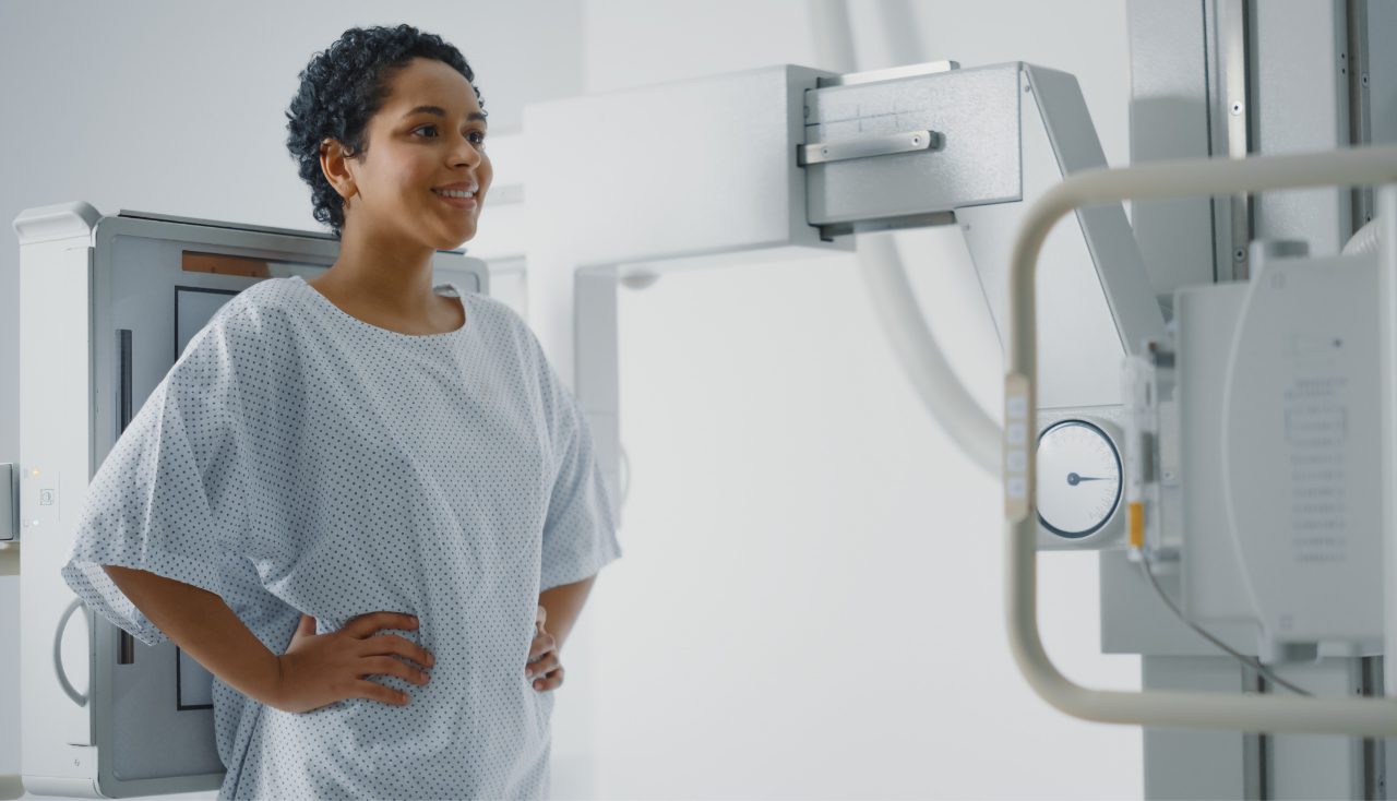 Hospital Radiology Room: Beautiful Smiling Latin Woman Standing next to X-Ray Machine While it Scans Chest Chest, Back, Lungs. Modern High-Tech Laboratory with Advanced Technical Equipment